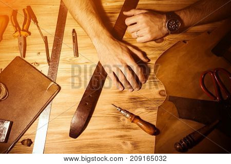 Working process of the leather belt in the leather workshop. Man holding hands on wooden table. Crafting tools on background. Tanner in old tannery. Close up men arm. Warm Light for text and design.