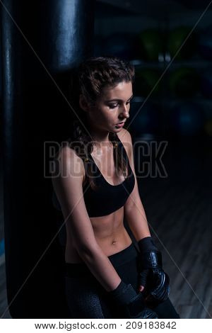 Upset young fighter boxer girl wearing boxing gloves standing near punching bag after loosing fight. She is sad and looking down.