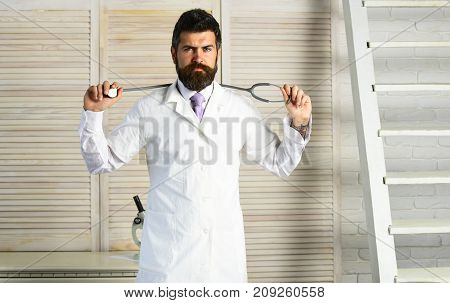 Man in surgical uniform with stethoscope on neck on wooden background. Healthcare and treatment concept. Physician with serious face ready to diagnose. Doctor in white medical coat holds equipment