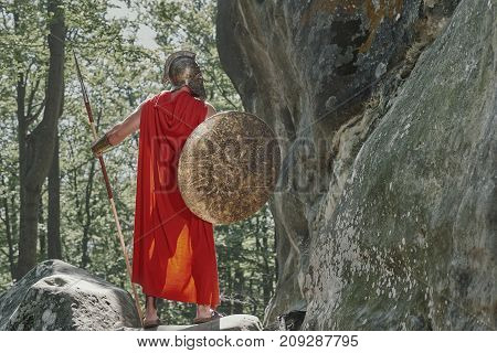 Rearview full length shot of a spartan warrior with a spear and a sword walking through the woods on a sunny day.