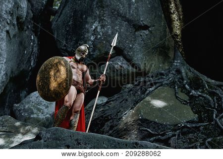 Portrait of a muscular powerful warrior in a helmet and cape posing dramatically on top of a rock in the woods copyspace.