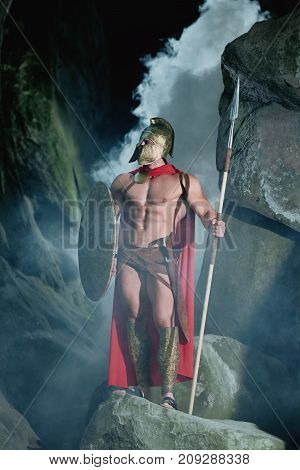 Vertical shot of a Spartan soldier walking through foggy dark forest with his shield and a spear in his hand.