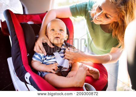 Young mother putting her little baby boy in the car seat, fastening seat belts.
