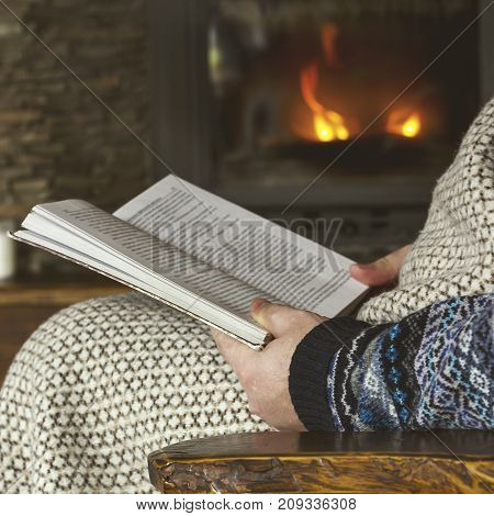 Man reading a book sitting in a rocking chair with knit plaid in front of a fireplace winter cozy concept soft toned image