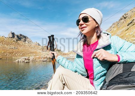 A tourist girl wearing sunglasses down jacket and hat with a backpack and mountain equipment with handles for tracking in her hands is sitting and relaxing looking at the beautiful view of the high mountain lake in the North Caucasus. Karachay-Cherkess Re