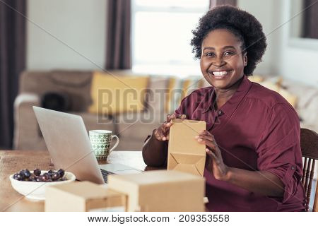 Smiling young African woman sitting at a table at home using a laptop and writing addresses on packages for her home based online business