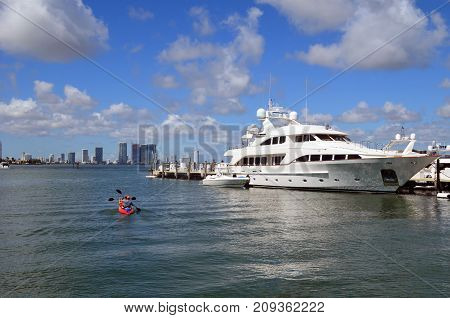 A couple in a rental kayak gliding by a luxurious white motor yachts moored at a marina on Miami Beach and in the distance Miami tall building skyline.