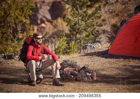Hicker with beard and sunglasses sitting near orange tent and hold a cup. Sunny spring daybreak in rocky mountains.