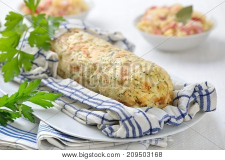 Uncut Bavarian bread dumpling cooked in a rolled up tea towel, sauerkraut with bacon in the background
