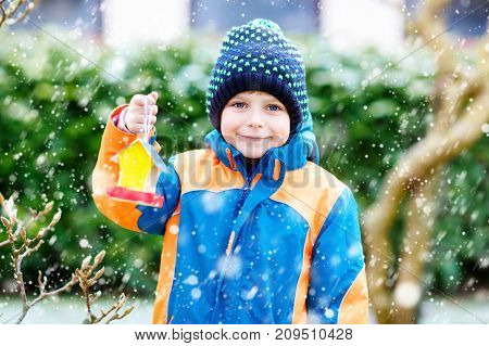 Little kid boy feeding birds in winter. Cute happy preschool child hanging colorful selfmade bird house on tree on frosty cold day. Preschooler in colorful wam clothes. Nature, empathy with animals.