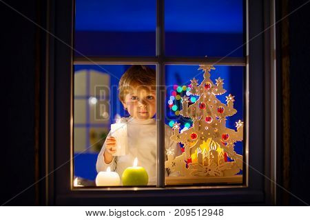 Beautiful little kid boy, standing by window at Christmas time and holding candle. Child waiting for Santa Claus. and gifts. With colorful lights from Christmas tree on background.