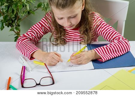 A  Junior Schoolgirl With Glasses Writes Something With Her Left Hand In The Notebook And Sits At Th