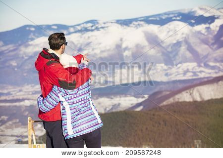 Rear view of a loving couple in fur hood jackets looking at snowed mountain range.
