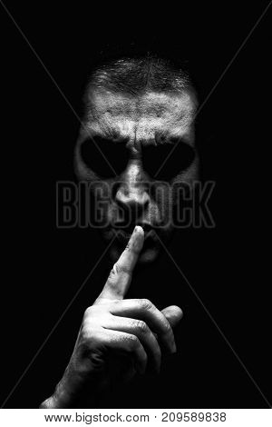 Angry mature man with an aggressive look making the silence sign in a threatening and creepy way. Black and White, black background. Concept for secret, threat, anger, rage, violence, danger, menace. 