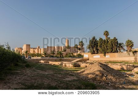 Stone walls of old Ribat fortress and minaret of the Grand Mosque through the lush palms of local garden. Ribat in Monastir is nearly 1200 years old in Monastir. Tunisia.