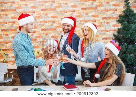 Attractive cheerful coworkers having fun and drinking at the Christmas event on a blurred office background. Business colleagues in red Santa hats.
