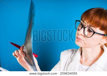 a nurse in big glasses with a serious face examines an X-ray and holds a pen in her hand, isolated on a blue background