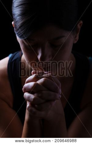 Low key portrait of a faithful woman praying, hands folded in worship to god, head down and eyes closed in religious fervor, on a black background. Concept for religion, faith, prayer and spirituality