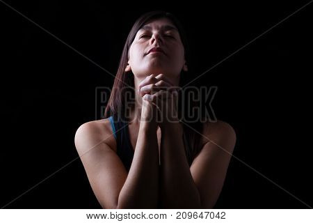 Close up of a faithful woman praying, hands folded in worship, head up and closed eyes in religious fervor, on a black background. Concept for religion, faith, prayer and spirituality.
