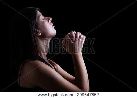 Close up of a faithful woman praying, hands folded in worship, head up and closed eyes in religious fervor, on a black background. Concept for religion, faith, prayer and spirituality.