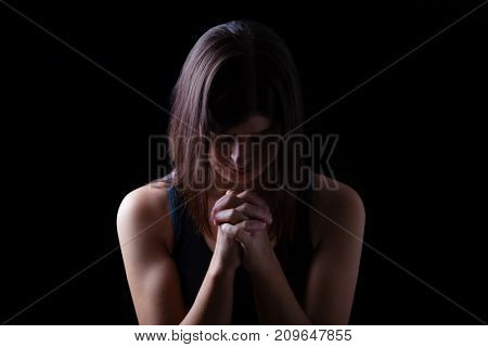 Faithful athletic woman praying, with hands folded in worship to god, head down and eyes closed in religious fervor, on low key black background. Concept for religion, faith, prayer and spirituality