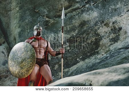 Shot of a medieval warrior with stunning athletic ripped body posing on a rock wearing helmet and a cape holding a shield and a sword ready for battle.