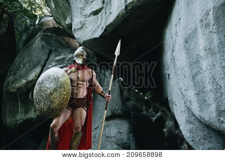 Portrait of a Spartan soldier walking through the forest near a rock carrying a spear and a shield.