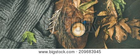 Autumn or Fall morning coffee concept. Flat-lay of knitted woolen grey sweater, wooden tray, mug of coffee and yellow fallen leaves over dark rustic wooden table background, top view, wide composition