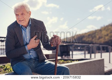 Worrying symptoms. Handsome white-haired man sitting on a concrete edge of a flower bed and suffering from a dull chest pain while wincing