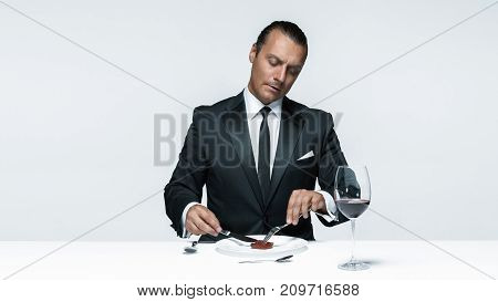 Portrait of a young man in image of maniak with knife, fork and and a piece of raw meat on a plate. Man in suit. The image in the style of Halloween