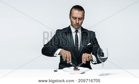 Portrait of a young man in image of maniak with knife, fork and and a piece of raw meat on a plate. Man in suit. The image in the style of Halloween