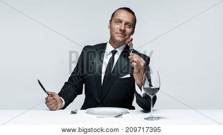 Portrait of a young man in image of maniak with knife, fork and and a piece of raw meat on a plate. Man in suit. The image in the style of Halloween