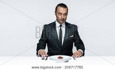 Portrait of a young man in image of maniak with knife, fork and and a piece of raw meat on a plate. Man in suit. The image in the style of Halloween