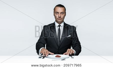 Portrait of a young man in image of maniak with knife, fork and and a piece of raw meat on a plate. Man in suit. The image in the style of Halloween