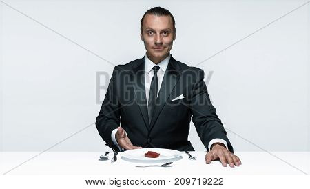 Portrait of a young man in image of maniak with knife, fork and and a piece of raw meat on a plate. Man in suit. The image in the style of Halloween