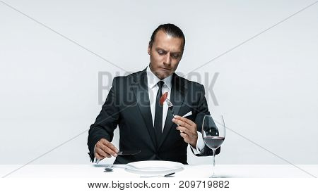 Portrait of a young man in image of maniak with knife, fork and and a piece of raw meat on a plate. Man in suit. The image in the style of Halloween