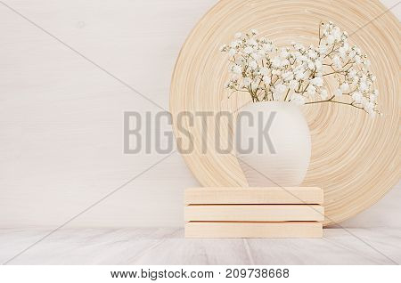 Soft home decor of beige bamboo dish and white small flowers in ceramic vase on white wood background. Interior.