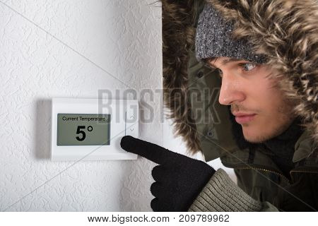 Young Man In Warm Clothing Pointing To Current Room Temperature In Digital Thermostat At Home