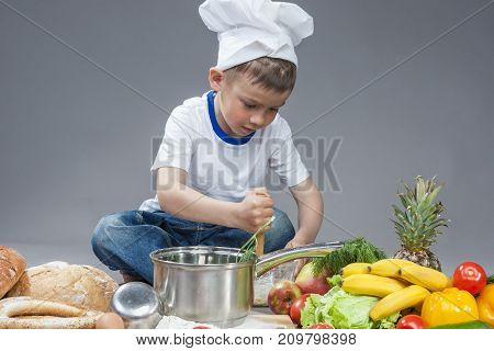 Cuisine Concepts. Portrait of Fascinated Caucasian Little Boy Working With Whisk In Cooking Hat. Surrounded by Fresh Fruits and Vegetables In Studio. Posing with Smiling Expression. Against Gray.Horizontal Image