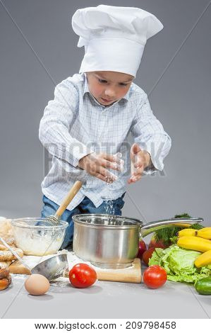 Cuisine Concepts.Portrait of Concentrated Caucasian Boy Working with Flour and Sieving it Over the Saucepan. Posing In Cooking Hat Surrounded by Fruits and Vegetables. Against Gray. Vertical Shot