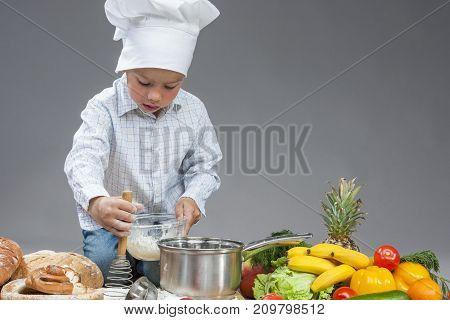 Portrait of Little Caucasian Boy Working With Whisk In Cooking Hat. Surrounded by Fresh Fruits and Vegetables In Studio. Posing with Smiling Expression. Against Gray.Horizontal Image Orientation
