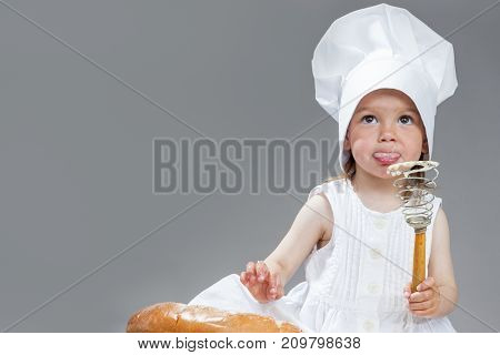 Funny Cooking Concepts. Portrait of Thinking Caucasian Little Girl in Cook Uniform Posing with Whisk Covered with Flour.Testing by Tongue. Against Gray. Horizontal Image