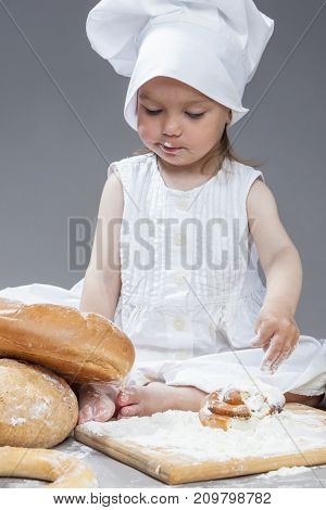 Cooking Ideas and Concepts. Portrait of Little Caucasian Girl in Cook Hat and Lips in Flour Sitting in Front of Fresh Buns. Against Gray.Vertical Image Composition