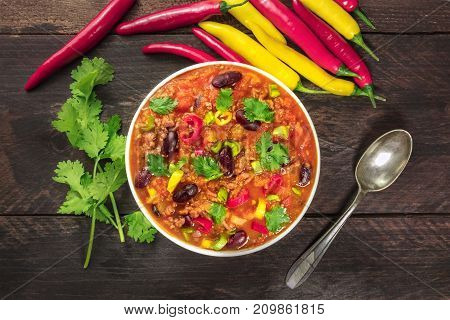An overhead photo of chili con carne, a traditional Mexican dish with red beans, cilantro leaves, ground meat, and chili peppers, on a dark rustic texture