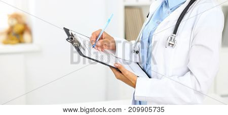 Close-up of a female doctor filling  up medical form at clipboard while standing straight in hospital.