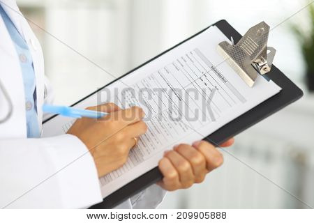 Close-up of a female doctor filling  up medical form at clipboard while standing straight in hospital.