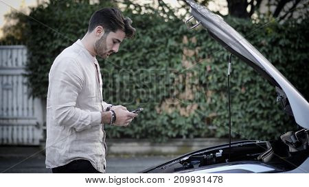 Handsome young man trying to repair a car engine, looking inside open bonnet then calling on cellphone, asking for help
