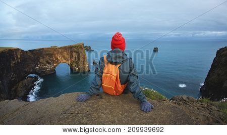 Girl in warm clothing, in red knitted hat and small orange backpack stands on the cliff on background of ocean, sky and Reynisfjara beach in Iceland. Popular tourist attraction. Unusual and gorgeous scene. Location Sudurland, cape Dyrholaey, coast of Icel