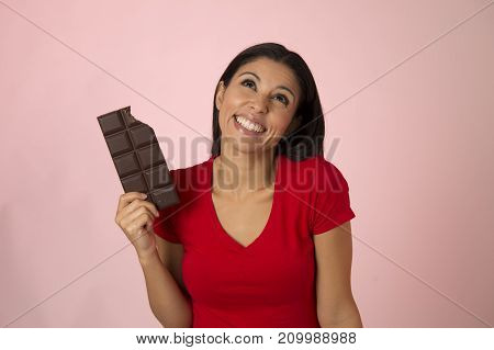 young attractive and happy hispanic woman in red top smiling excited eating chocolate bar isolated on pink background in diet calories and sugar addiction concept and sweet temptation concept