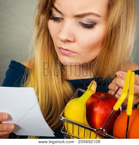 Worried woman holding shopping basket with fruits looking at bill receipt being scared of huge prices